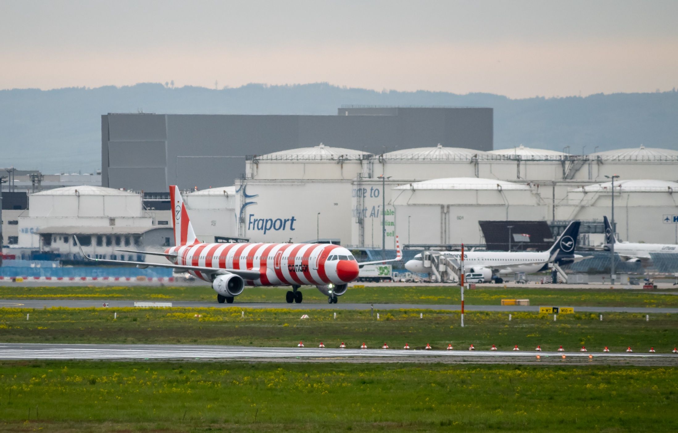 Condor-Flugzeug mit rot-weißem Streifenmuster am Frankfurter Flughafen, Fraport-Anlagen und Lufthansa-Flugzeuge im Hintergrund.