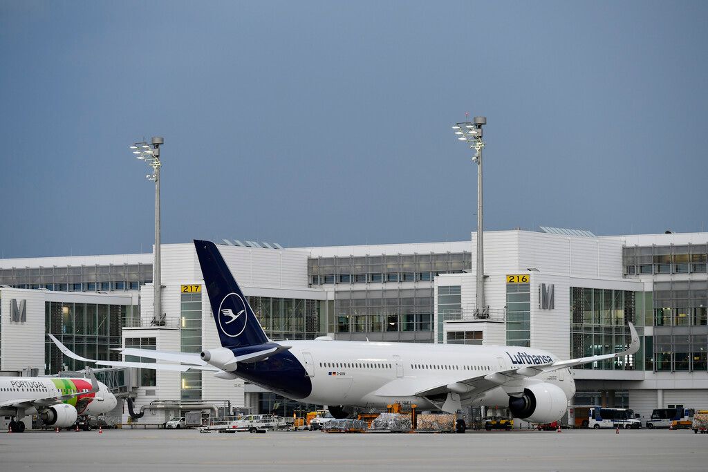 A Lufthansa aircraft parked at an airport terminal gate, with a TAP Air Portugal plane visible in the background.
