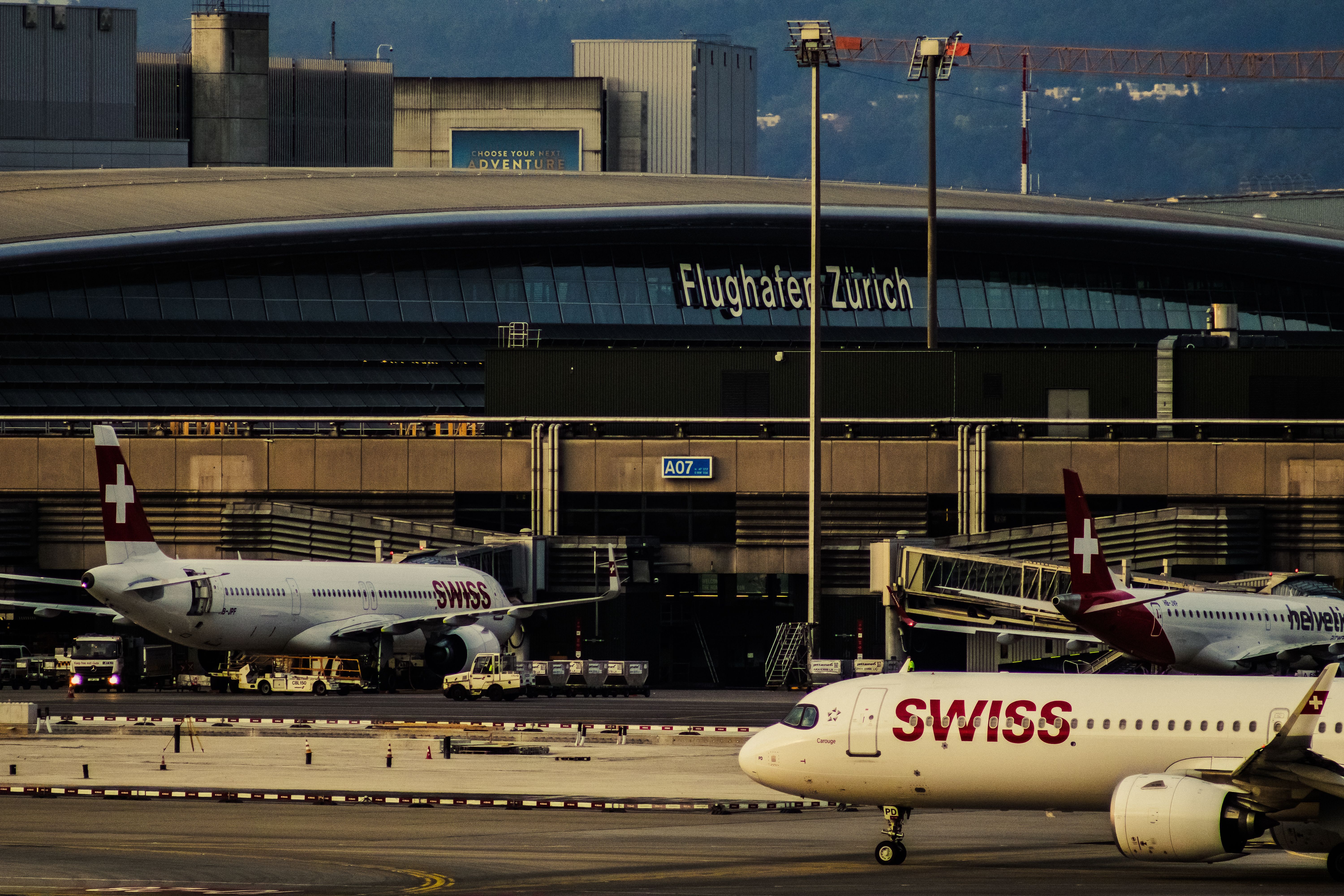Zwei SWISS-Flugzeuge, darunter 'Carouge', am Rollfeld des Flughafen Zürich, mit einem "CHOOSE YOUR NEXT ADVENTURE"-Schild am Terminal.