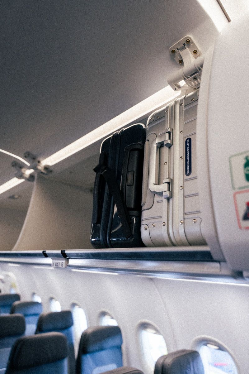 Two suitcases stored in an airplane's overhead bin above passenger seats, illustrating the cost of carry-on space in Economy Basic fares.