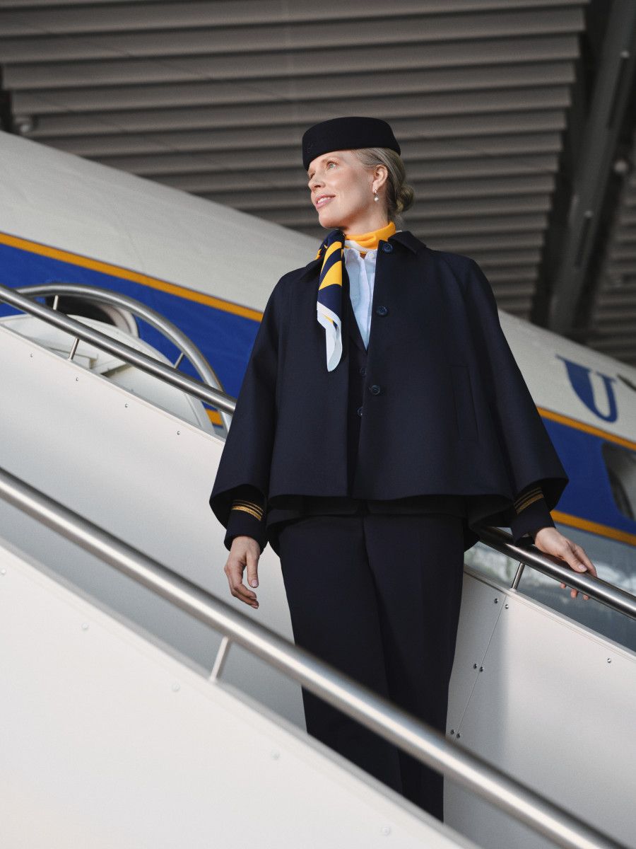 A flight attendant in a dark blue cape uniform standing on airplane stairs, looking away from the camera.
