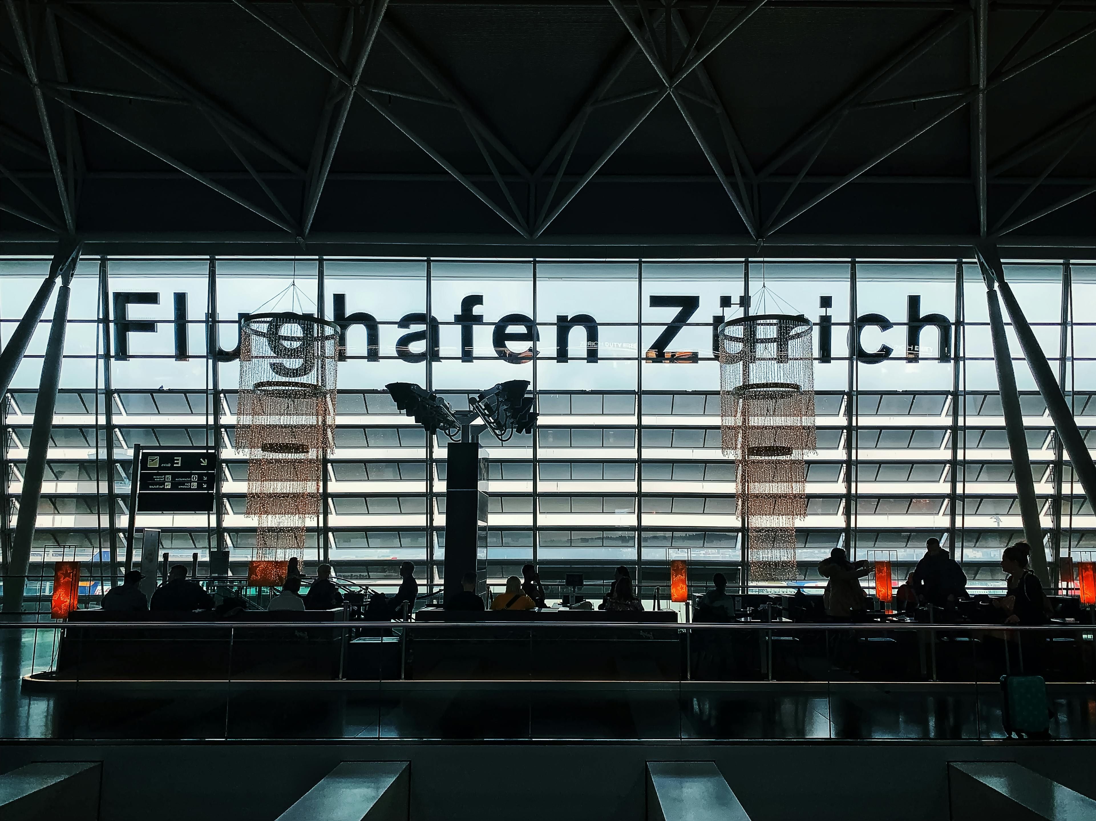 Innenansicht Flughafen Zürich: Wartende unter dem Schriftzug "Flughafen Zürich" und dekorativen Kronleuchtern.