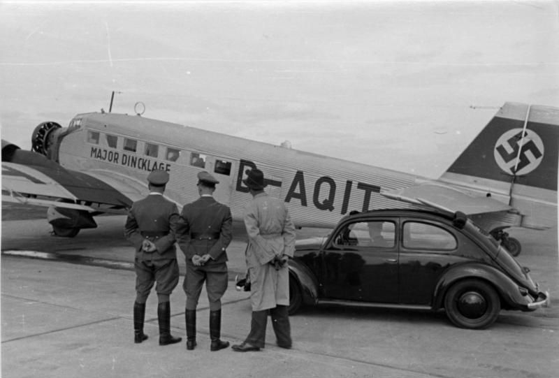 Three men stand before a Ju 52 aircraft marked 'Major Dincklage' and a Volkswagen Beetle at an airfield during the Third Reich era.