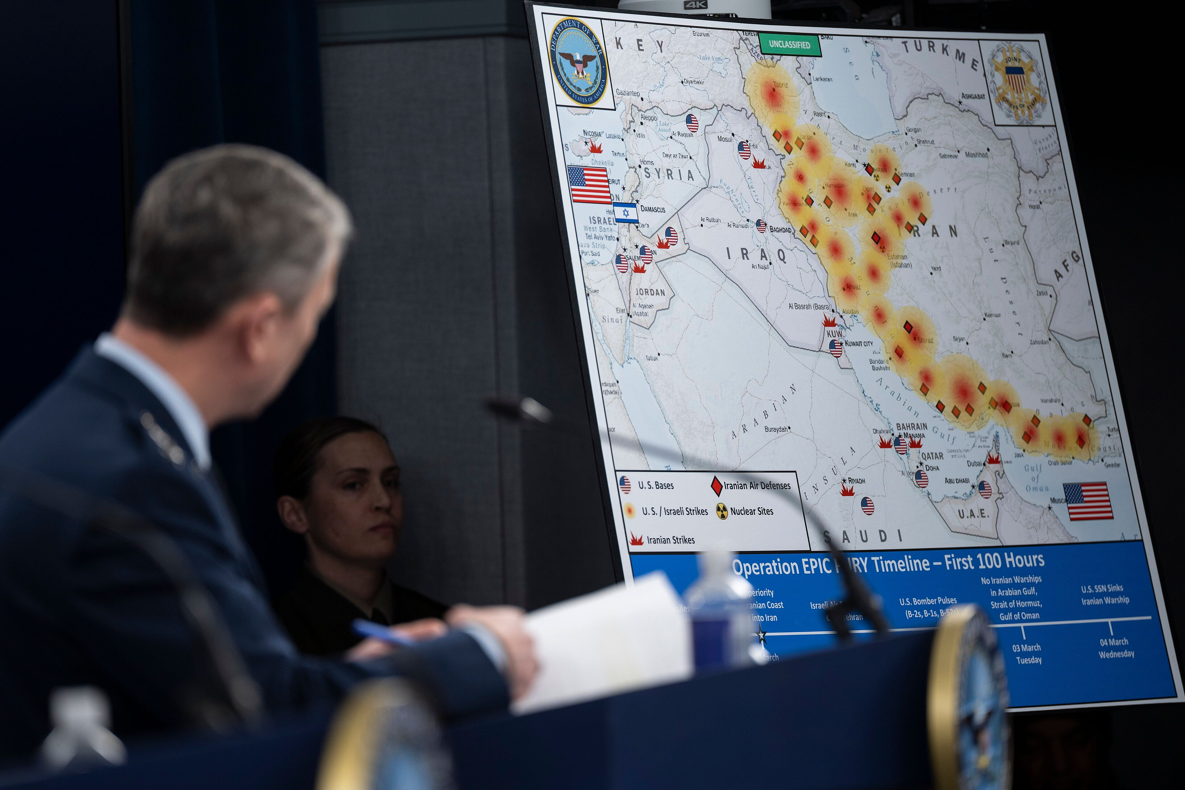 Briefing map showing strikes in the Middle East with US and Iranian flags, with officials in foreground.