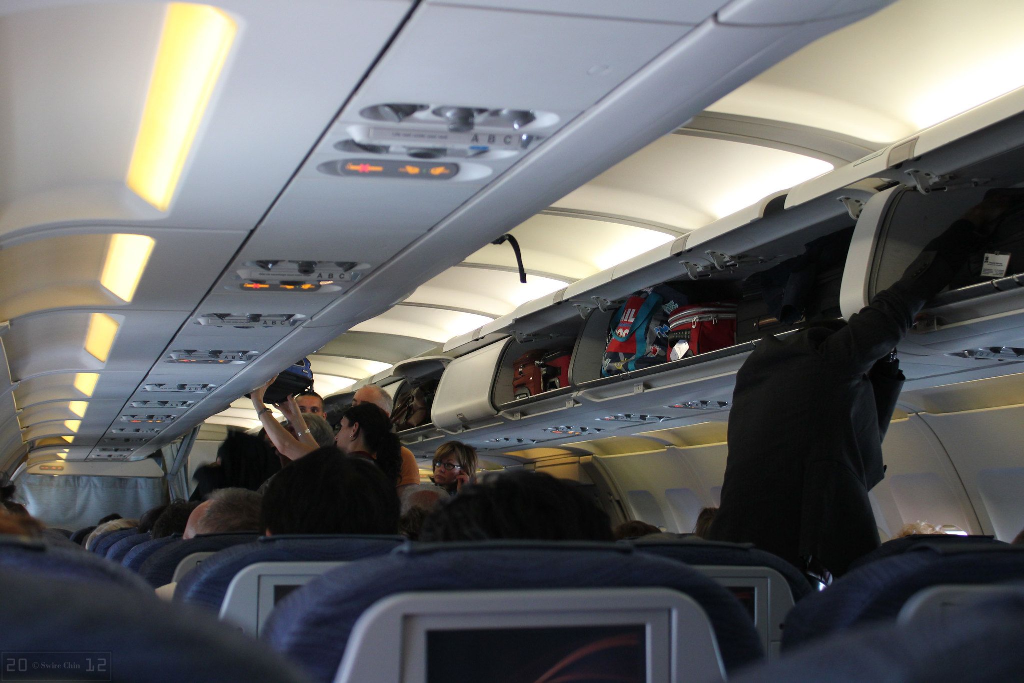 Passengers struggling to fit carry-on luggage into crowded overhead bins on a commercial airplane.