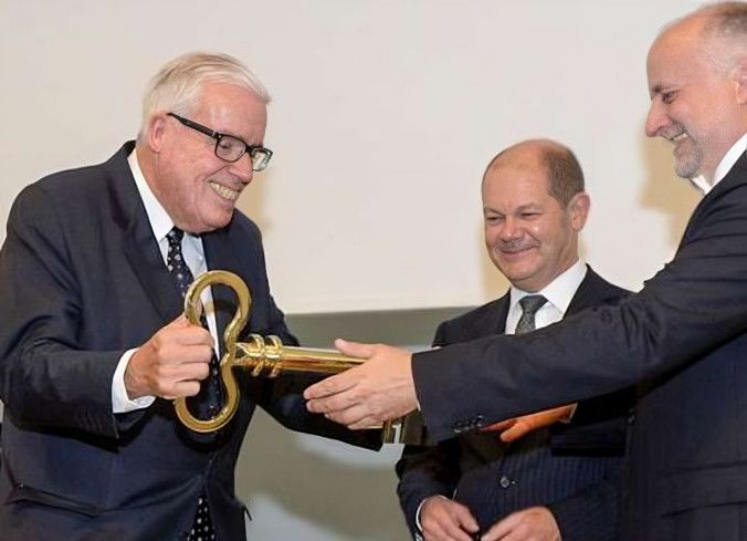 Klaus-Michael Kühne receives a large, golden key from another man, with Olaf Scholz in the background.