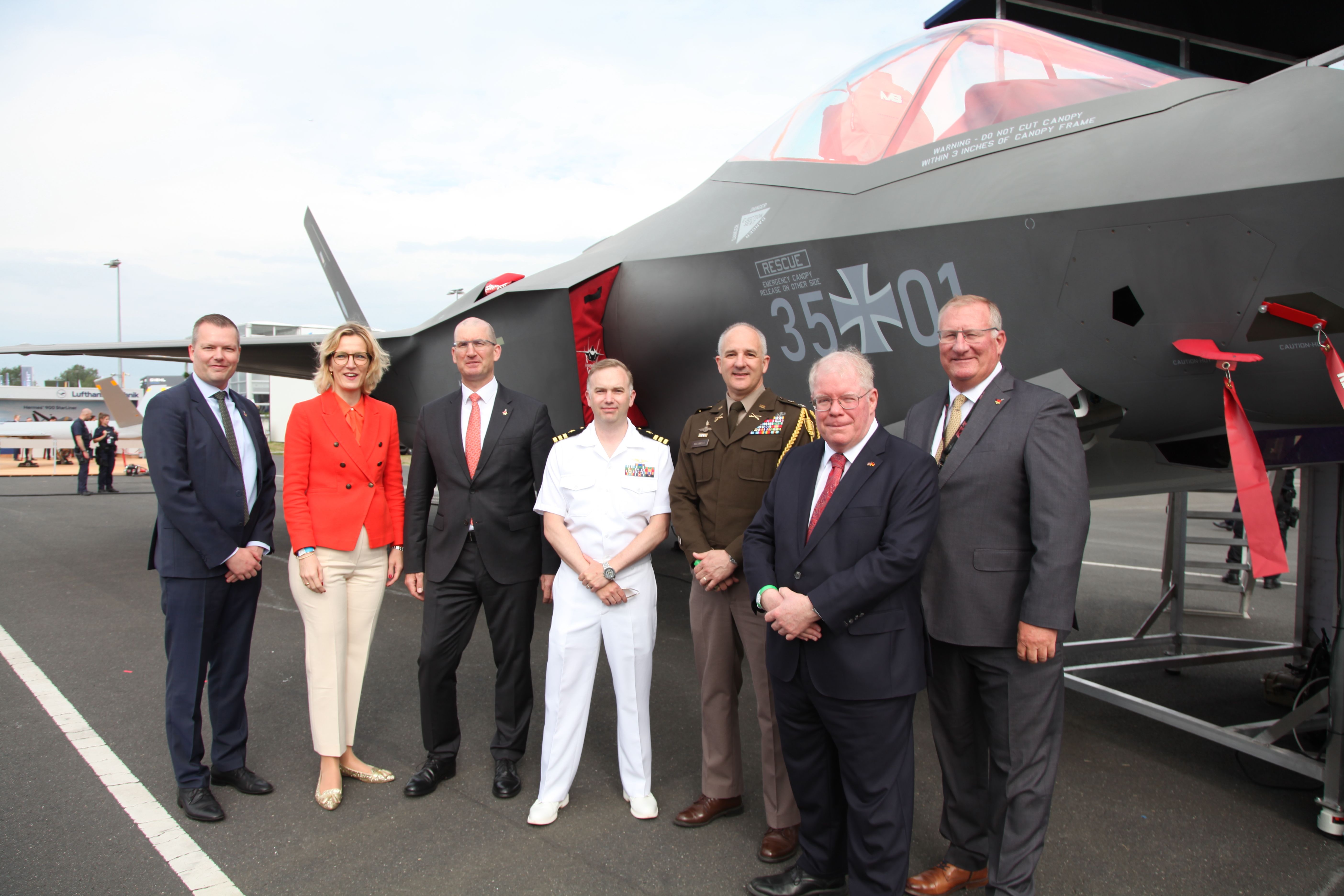 Military officials and executives pose before a German F-35 fighter jet at an airshow.