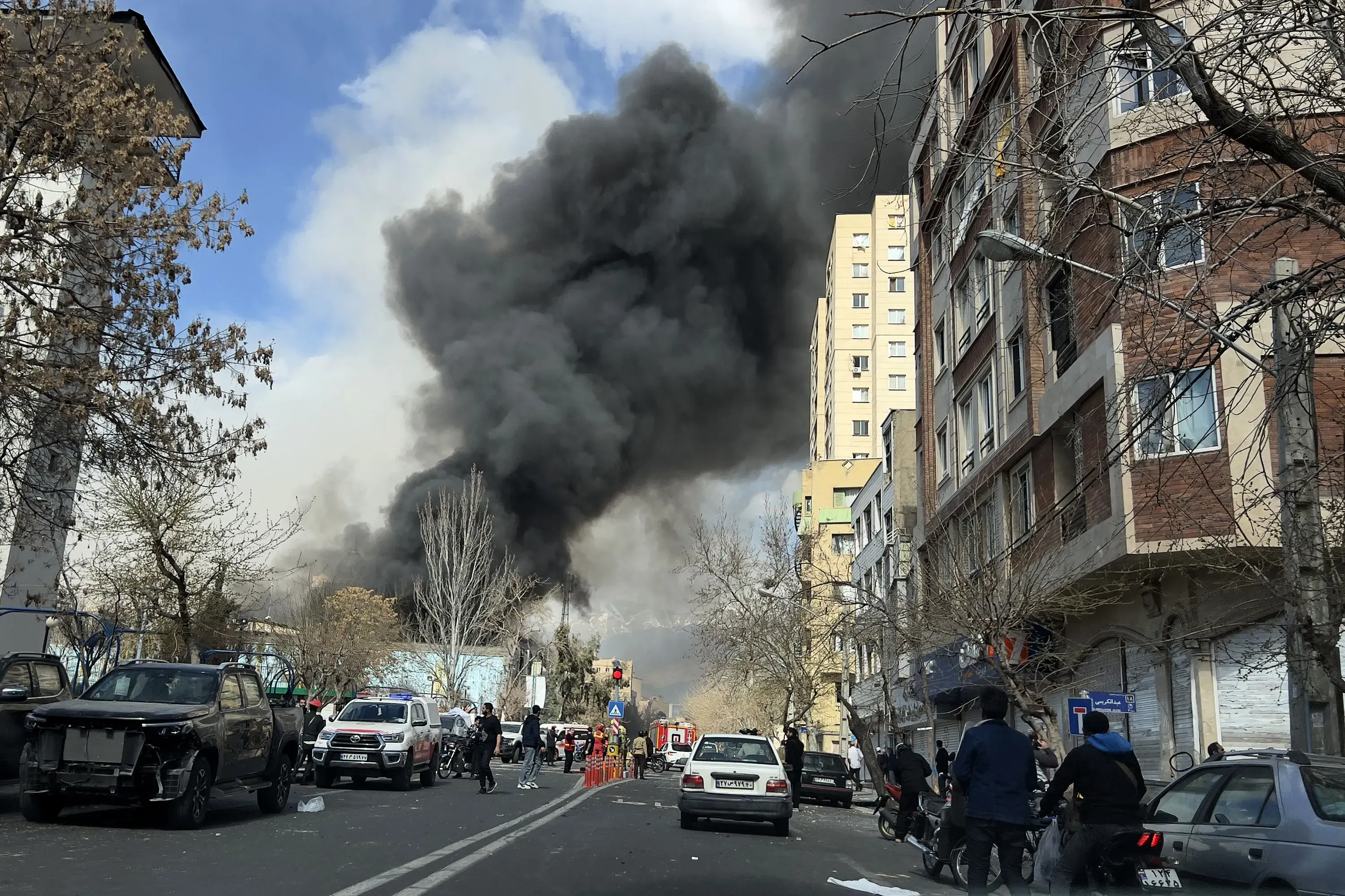Tehran street scene with thick black smoke in background. Emergency vehicles present.