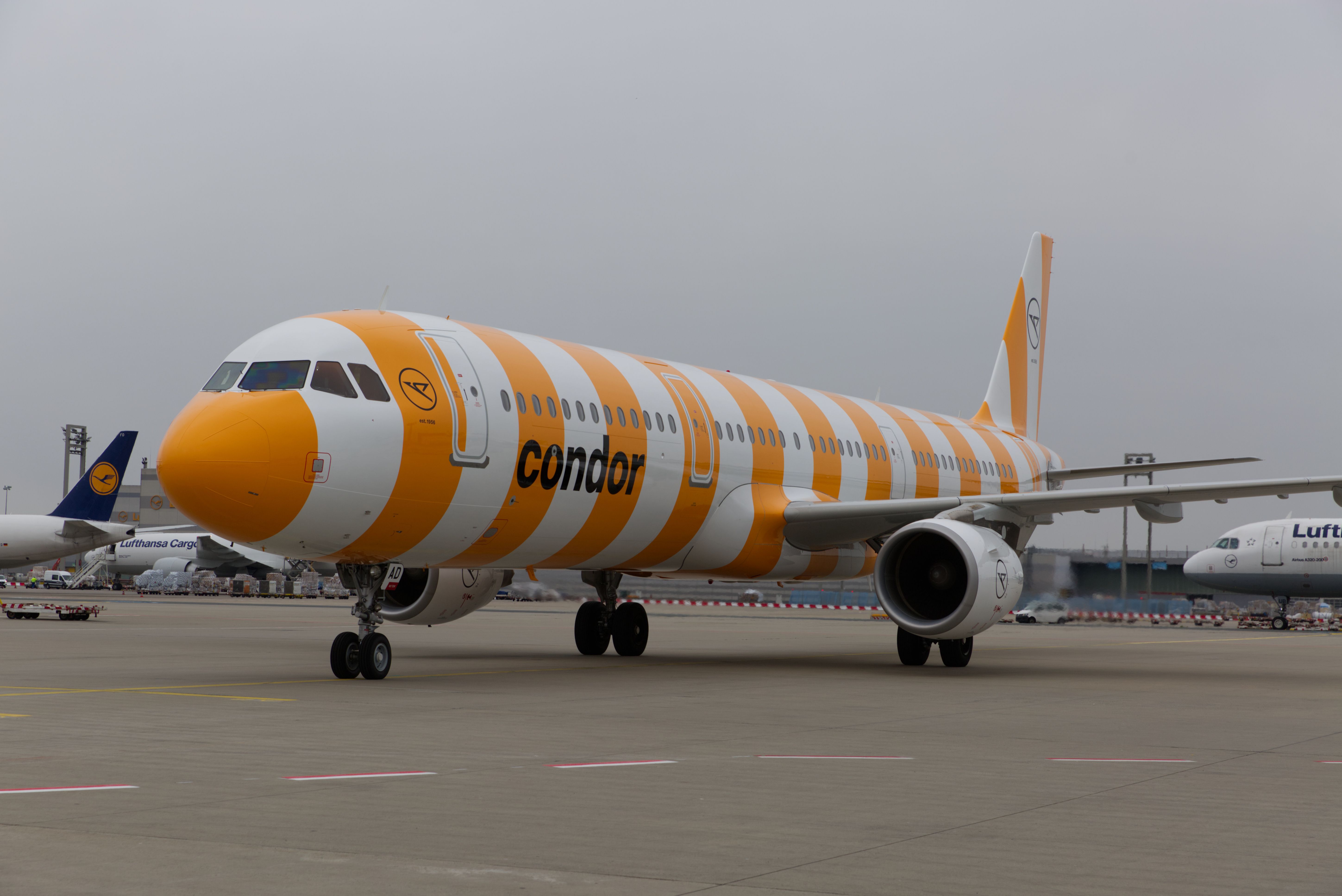 Condor Airbus A321 (D-AIAD) in gelb-weiß gestreifter 'Sunshine'-Lackierung auf dem Vorfeld, mit Lufthansa-Flugzeugen im Hintergrund.