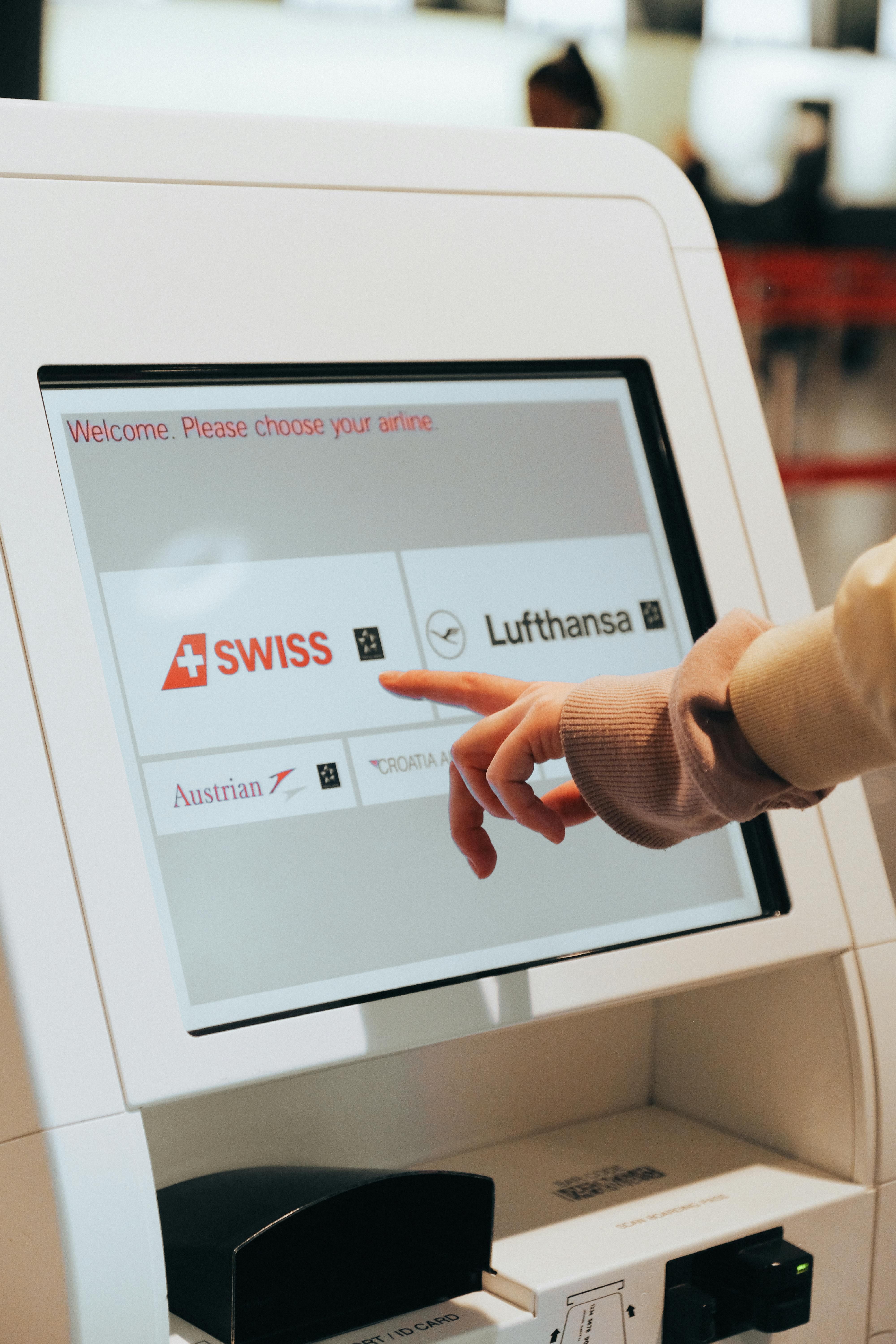Flughafen-Kiosk: Hand wählt Fluggesellschaft (Swiss, Lufthansa, Austrian, Croatia Airlines) auf Touchscreen.